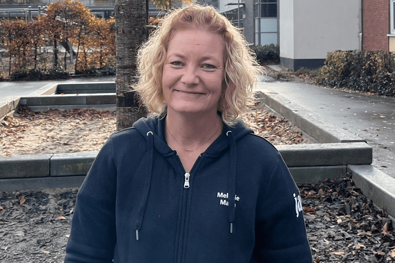 Frau mit blonden Locken in schwarzer Jacke steht in einem herbstlichen Umfeld mit Laub. Im Hintergrund Gebäude sichtbar.