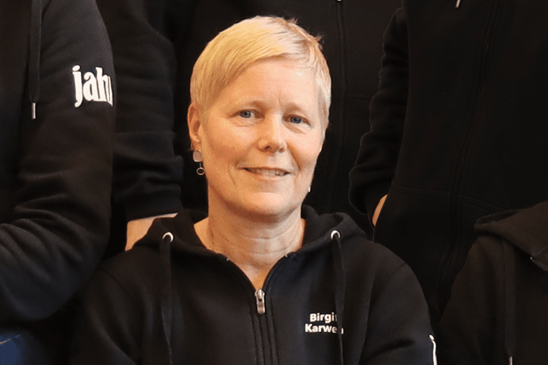 Frau mit kurzen blonden Haaren sitzt l&auml;chelnd auf einer Couch, tr&auml;gt einen schwarzen Kapuzenpullover und Jeans.