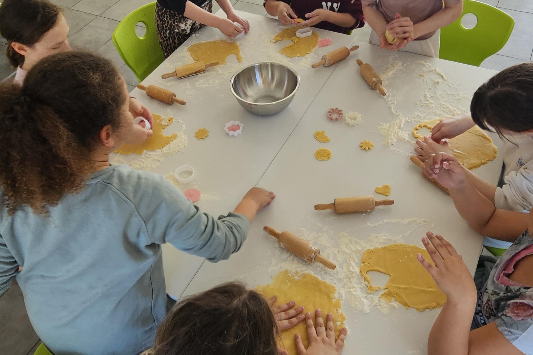 Gruppe von Kindern beim Pl&auml;tzchenbacken an einem Tisch mit Teig und Backutensilien in einer hellen K&uuml;che.