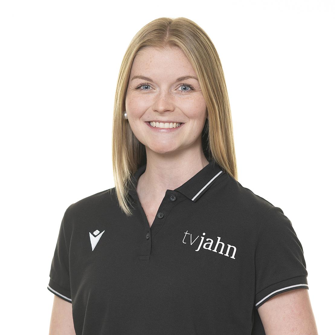Portr&auml;t einer l&auml;chelnden Frau mit blonden Haaren in einem schwarzen Polohemd mit Logo von tvjahn. Hintergrund hell.