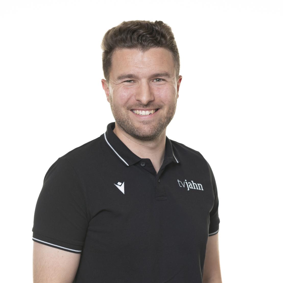 Junger Mann mit kurzem Haar l&auml;chelt im schwarzen Polo-Shirt vor hellem Hintergrund.