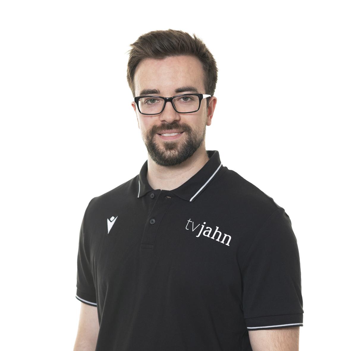 Porträt eines Mannes mit Brille, der ein schwarzes Poloshirt mit dem Aufdruck „tvjahn“ trägt, vor einem weißen Hintergrund.