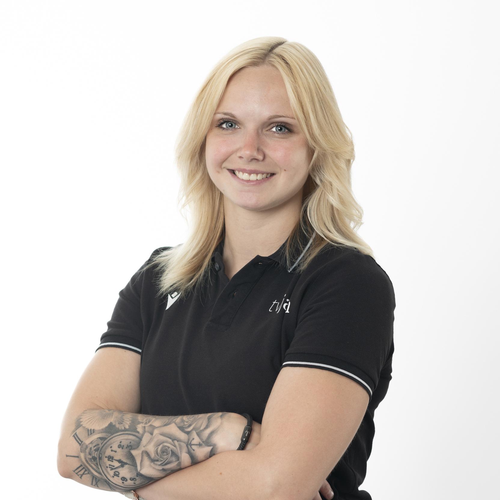 Portr&auml;t einer l&auml;chelnden Person mit blonden Haaren, die in einem schwarzen Poloshirt posiert und einen Arm mit Tattoos zeigt.