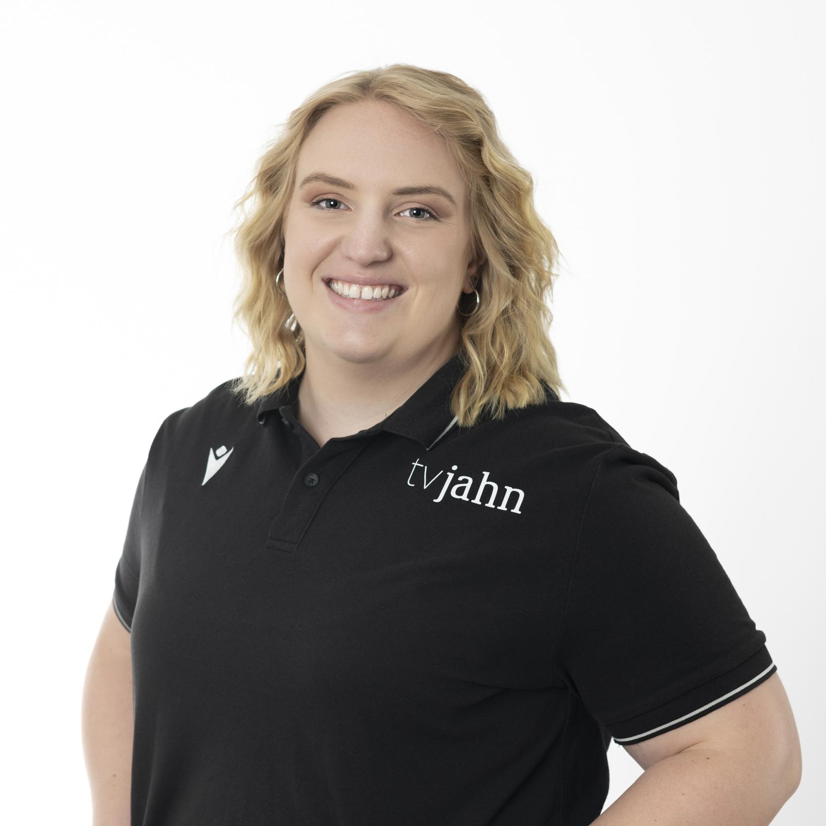 Portr&auml;t einer l&auml;chelnden Person mit blonden, welligen Haaren, die ein schwarzes Poloshirt mit Logo tr&auml;gt.