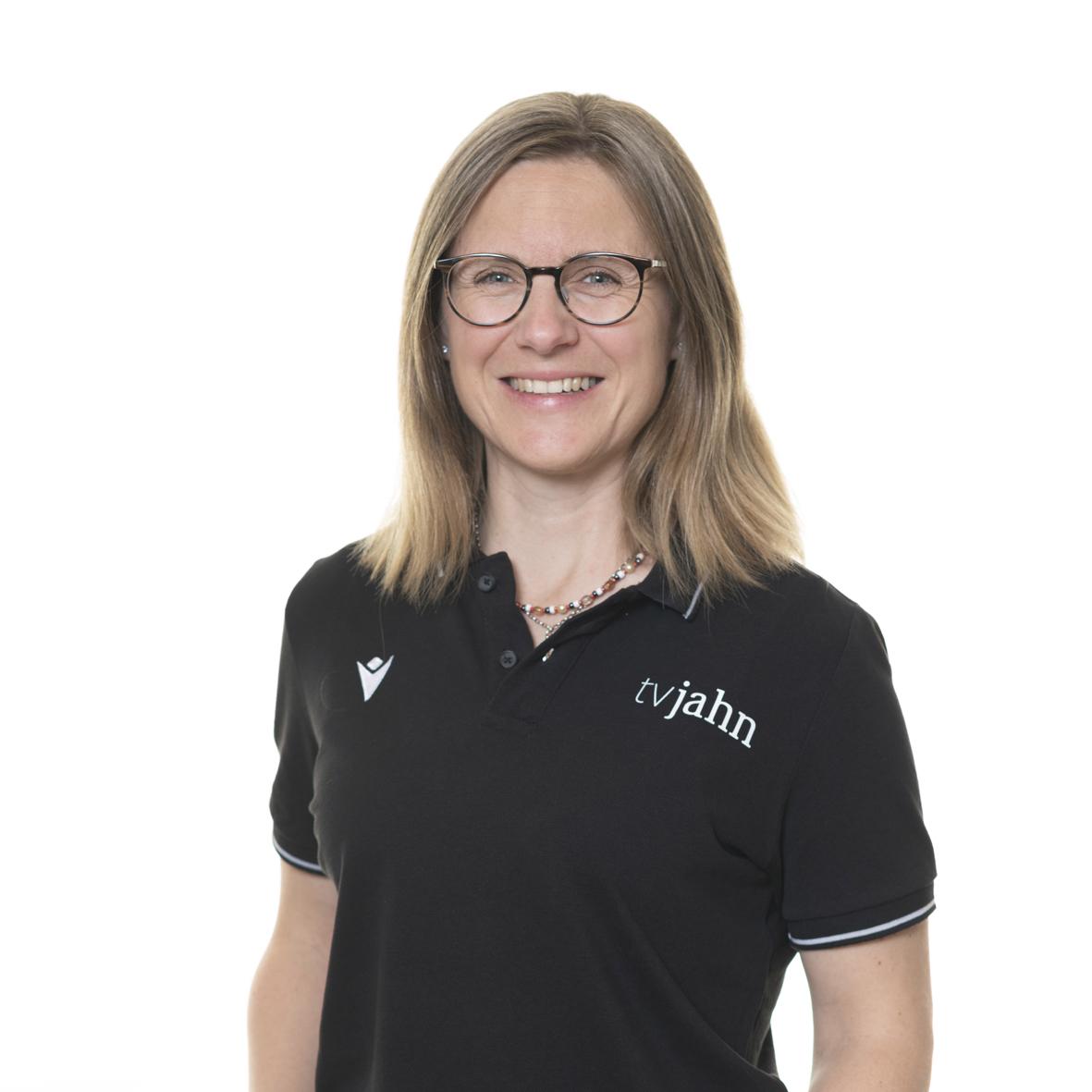 Portrait einer l&auml;chelnden Frau mit br&uuml;nettem Haar, Brille und schwarzem Poloshirt mit Logo von tvjahn.