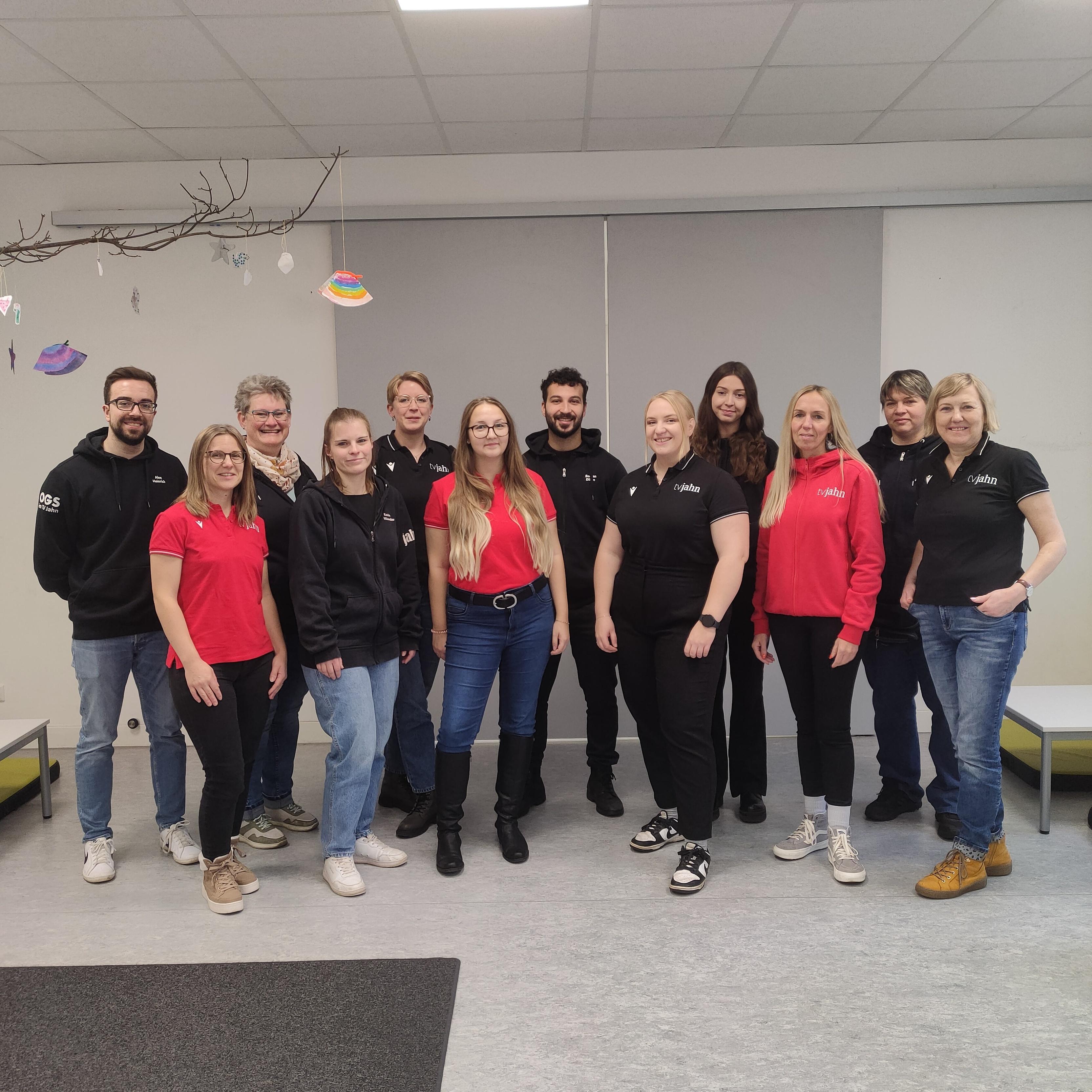 Gruppenfoto von zwölf Personen, die in einheitlicher Kleidung in einem hellen Raum mit Tischen und Dekoration stehen.