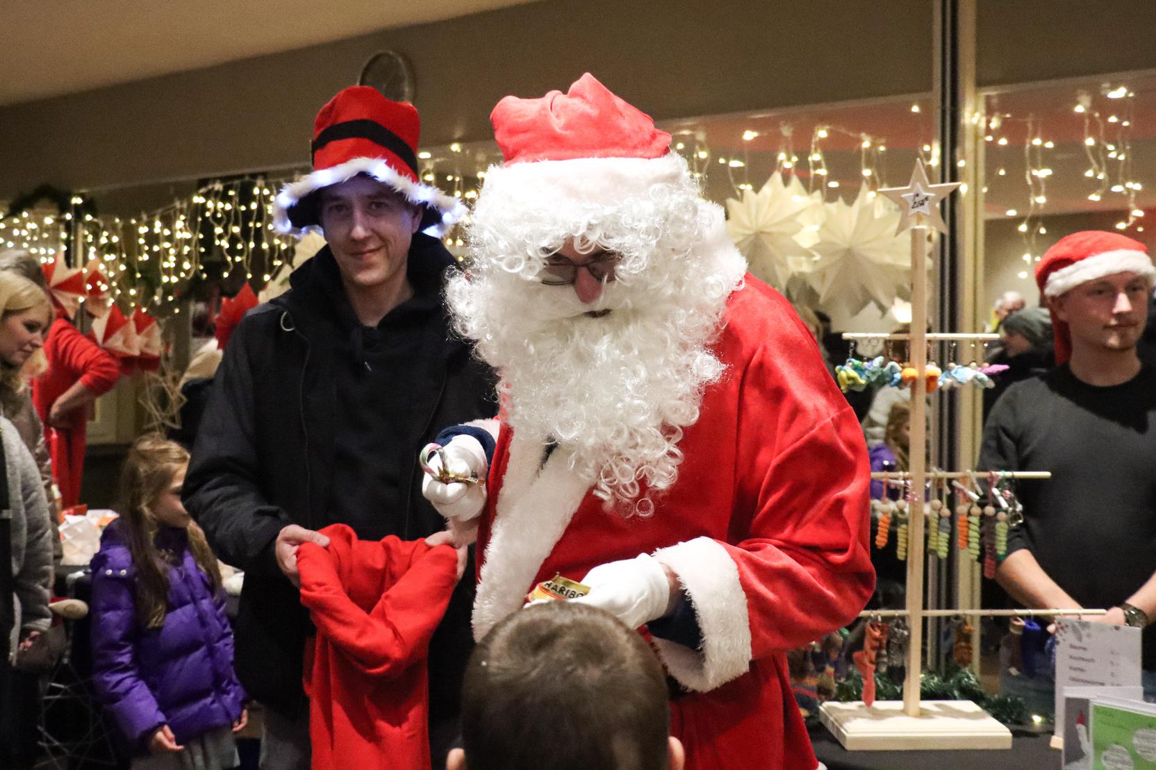 Weihnachtsmann verteilt Geschenke an Kinder, umgeben von festlich gekleideten Erwachsenen und Weihnachtsdekoration.