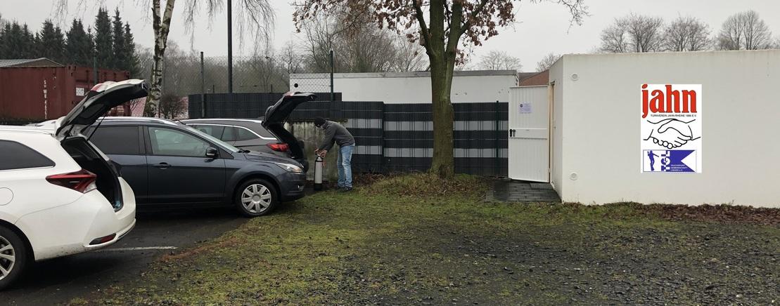 Person l&auml;dt Gegenst&auml;nde aus dem Kofferraum eines Autos auf einem Parkplatz vor einer Lagerhalle mit Firmenlogo.