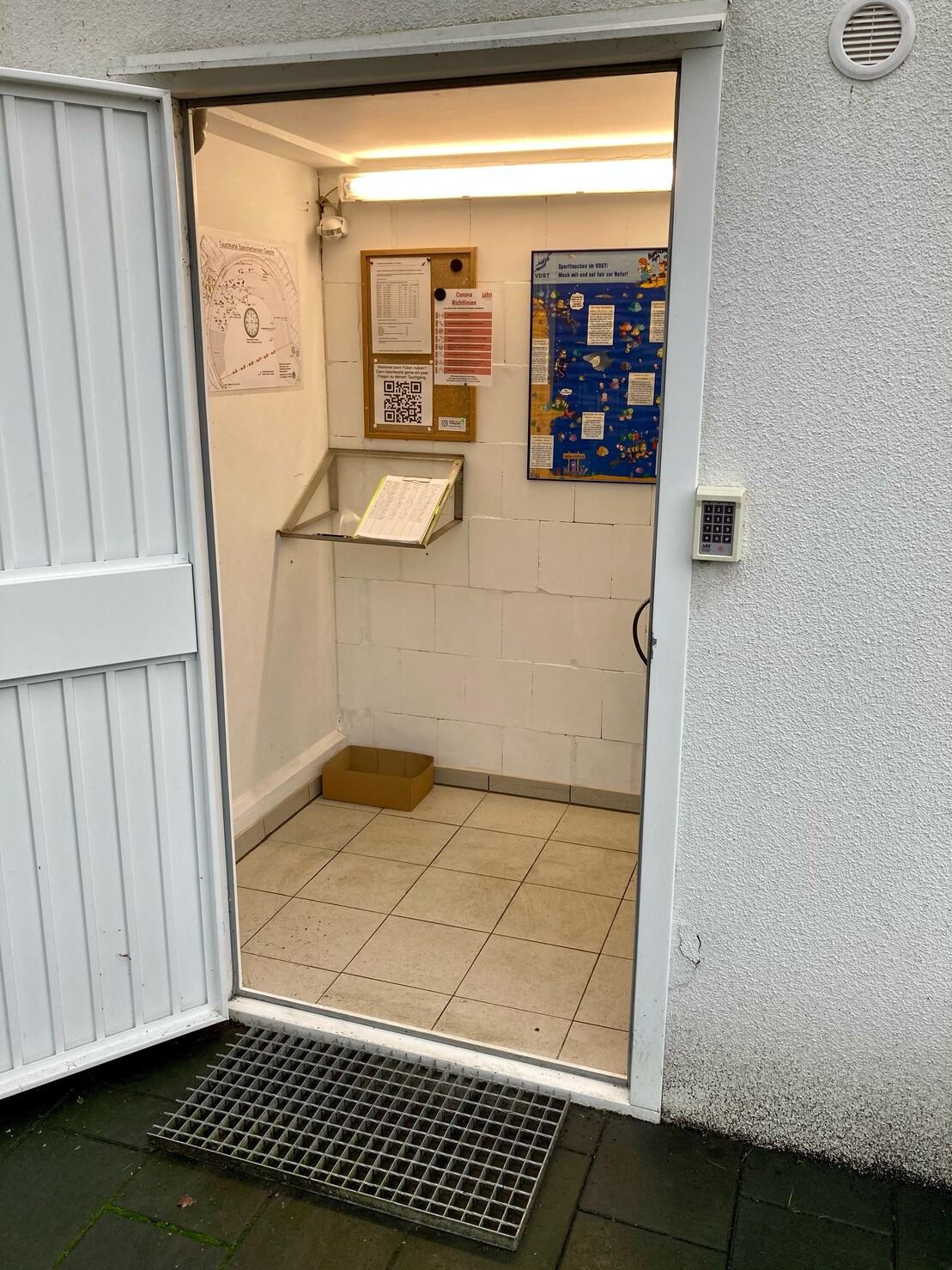 Eingang zu einem kleinen Raum mit informativen Aush&auml;ngen, einer Ablage und einem Karton auf dem Boden.