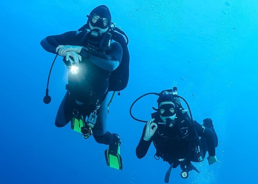 Zwei Taucher mit Ger&auml;ten und Taschenlampen schweben unter Wasser in blauer Umgebung.