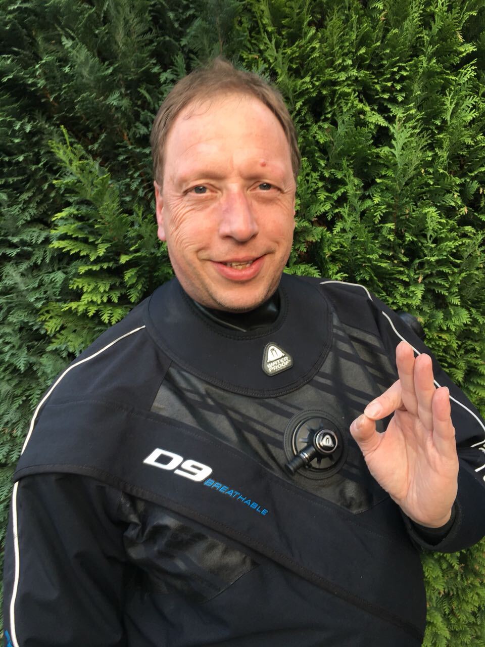Mann in einem Tauchanzug steht vor gr&uuml;ner Hecke und zeigt ein OK-Zeichen mit der rechten Hand.