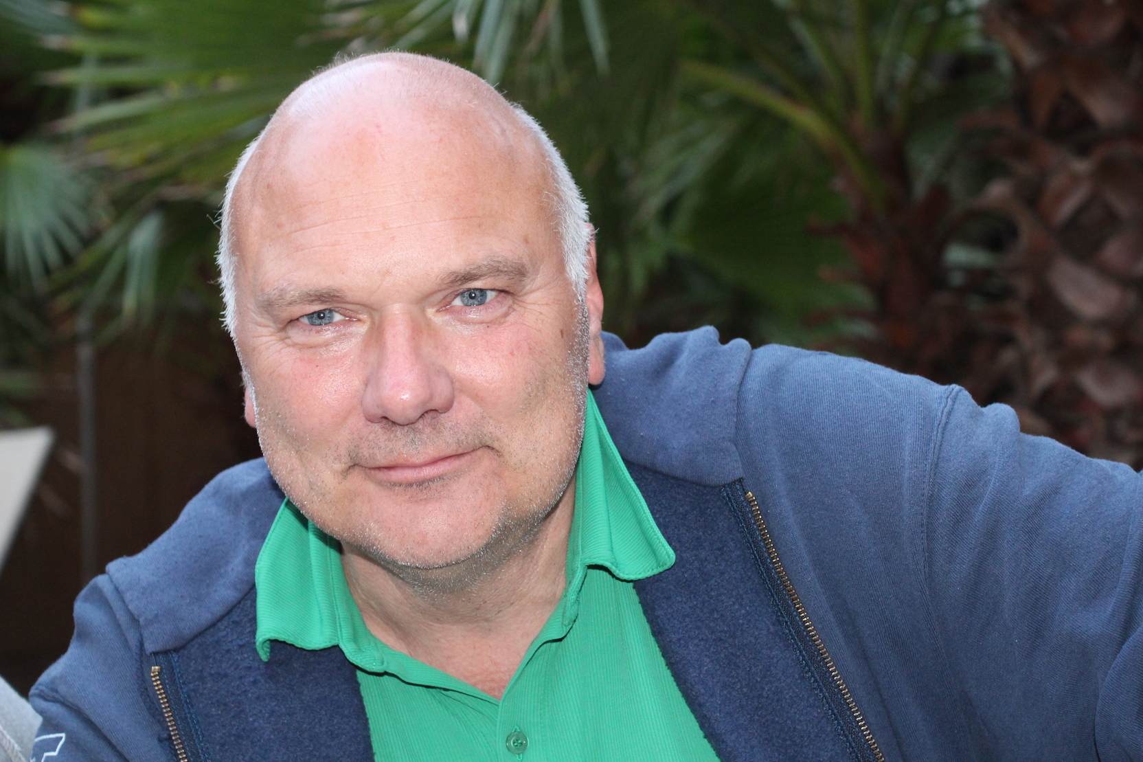 Mann mit Glatze und blauen Augen, tr&auml;gt ein gr&uuml;nes Hemd und einen blauen Pullover, vor einem Pflanzenhintergrund.