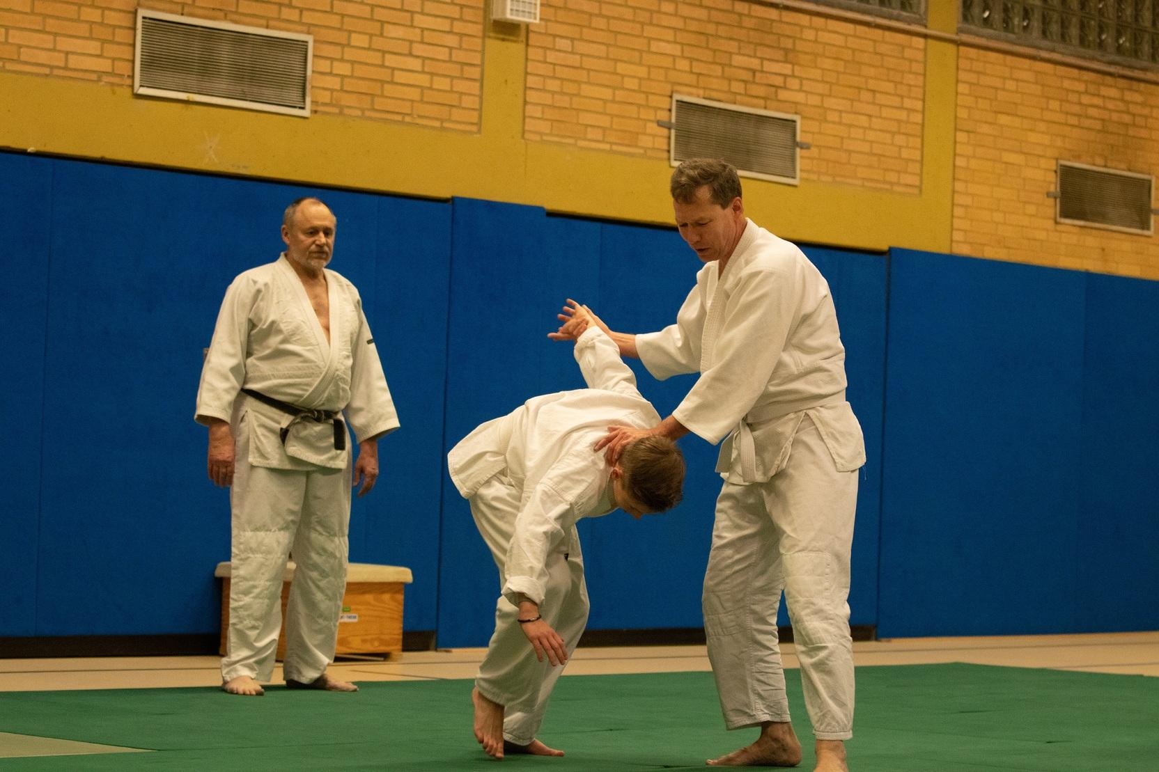Zwei Judoka f&uuml;hren eine Technik&uuml;bung auf einer Matte in einem Trainingsraum durch, w&auml;hrend ein Trainer zuschaut.