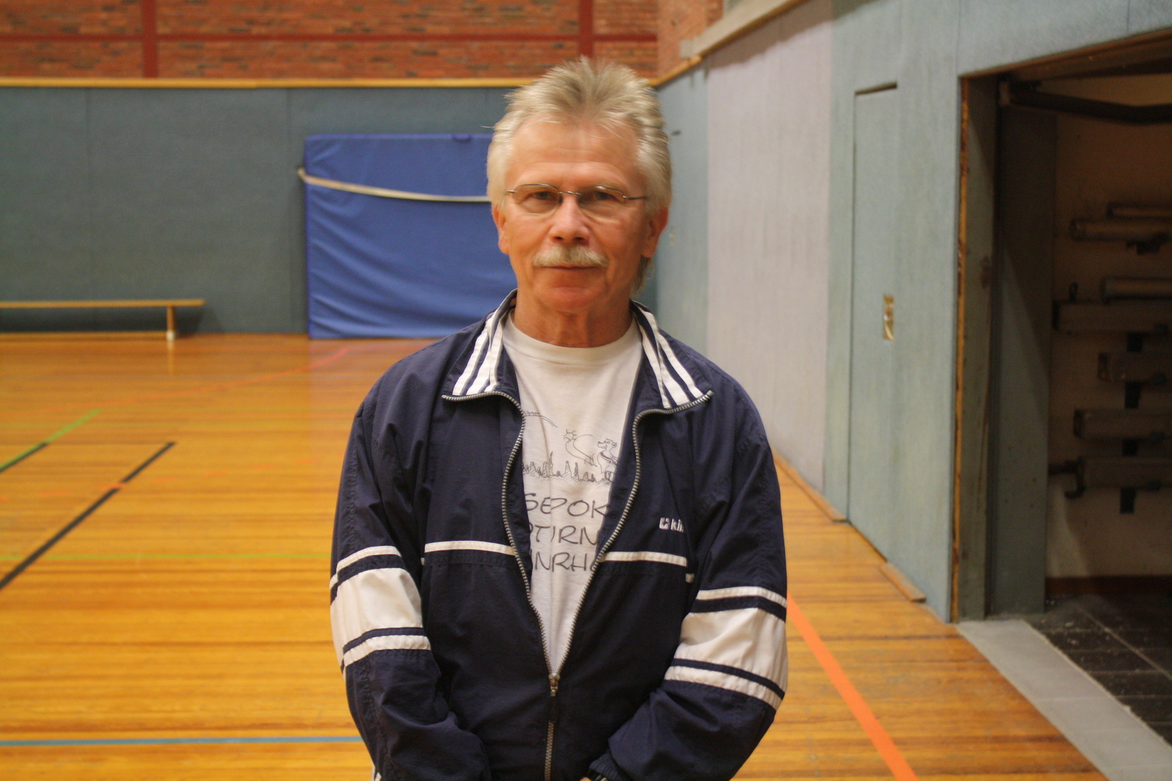 Ein &auml;lterer Mann in Sportkleidung steht in einer Halle mit Holzboden und Wand mit blauer Plane im Hintergrund.