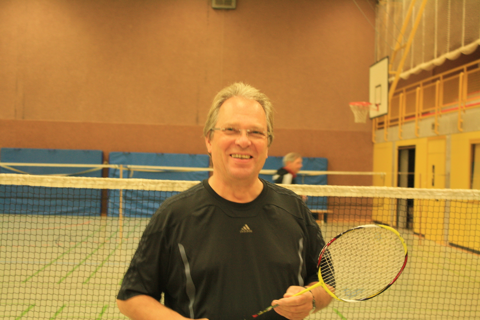 Mann mit Badmintonschl&auml;ger l&auml;chelt in einer Sporthalle vor einem Badmintonfeld.
