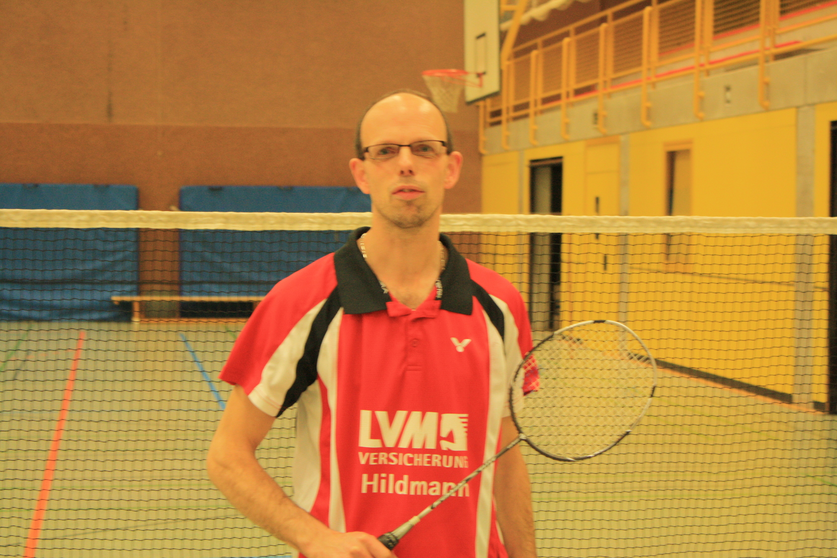 Mann mit Brille und Badminton-Schl&auml;ger in Sportkleidung steht vor einem Badmintonfeld.