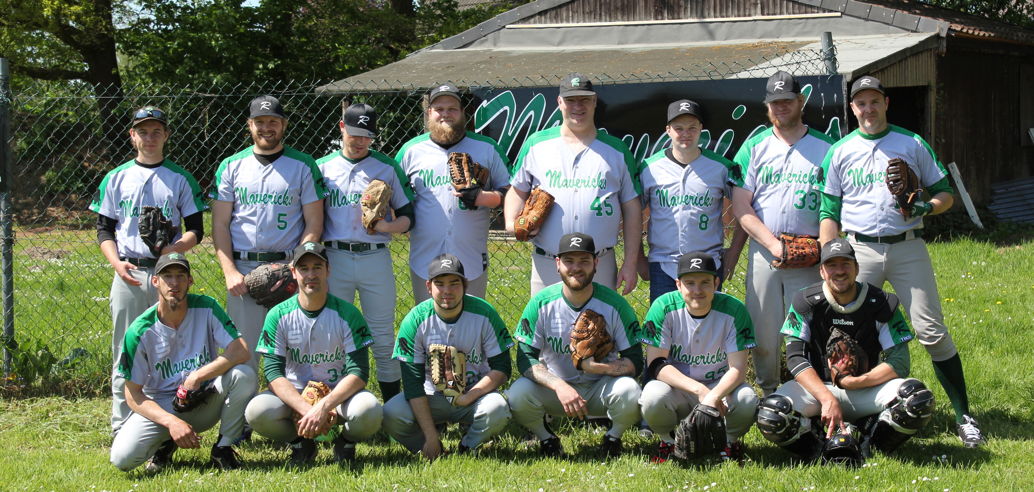 Mannschaftsbild von 13 Baseballspielern in gr&uuml;nen Trikots, auf einer Wiese vor einer Scheune, mit Handschuhen und -schl&auml;gern.