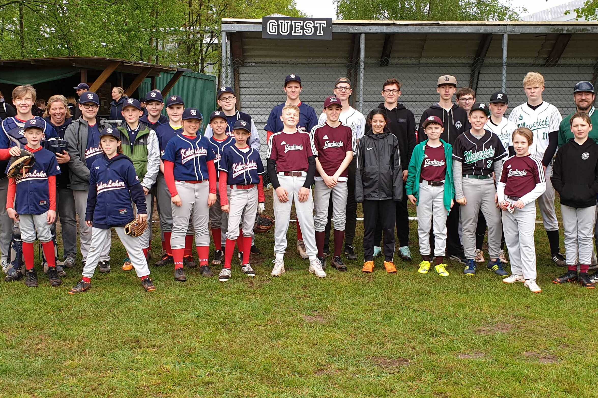 Gruppenfoto von Baseballspielern in Uniformen auf einem Sportplatz, mit B&auml;umen und einem Geb&auml;ude im Hintergrund.