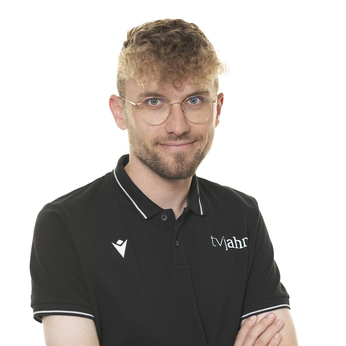 Junger Mann mit lockigem Haar und Brille, trägt ein schwarzes Polo-Shirt mit dem Logo "tvjahr".
