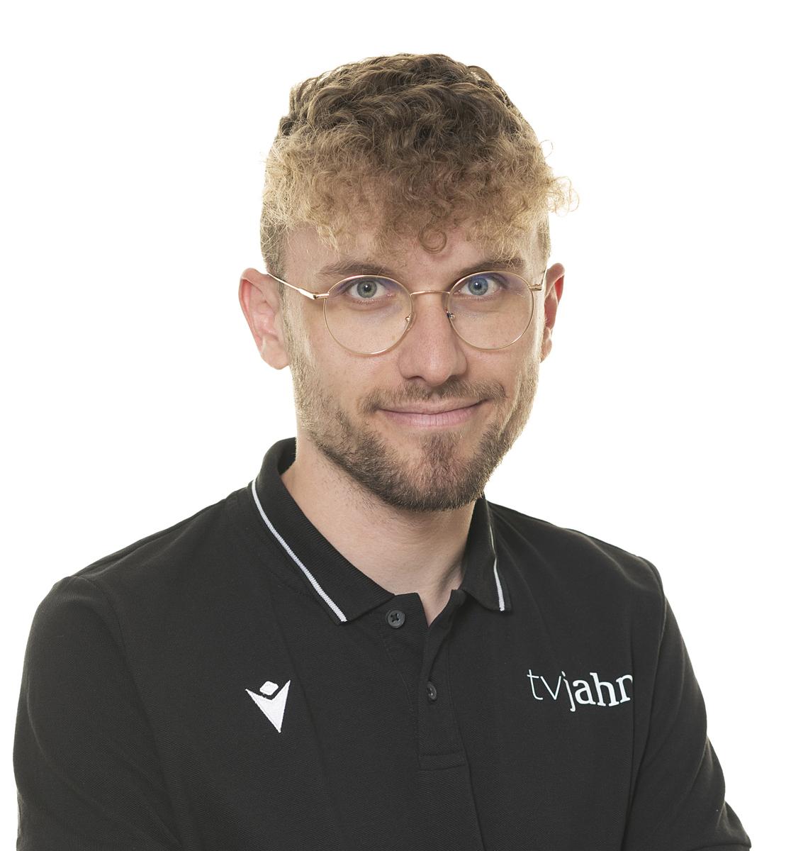 Junger Mann mit lockigem Haar und Brille, tr&auml;gt ein schwarzes Polo-Shirt mit dem Logo "tvjahr".