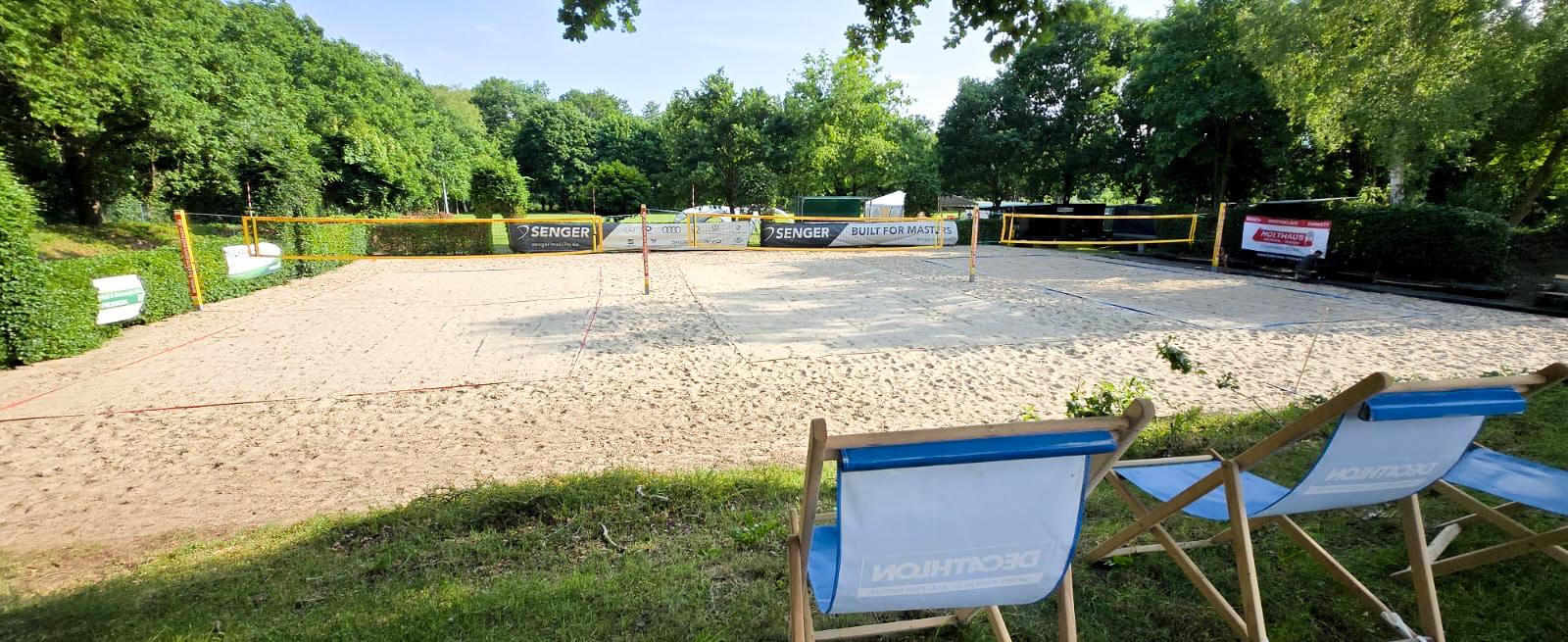 Sandvolleyballfeld umgeben von Bäumen, mit zwei Klappstühlen im Vordergrund und zahlreichen Werbebannern.