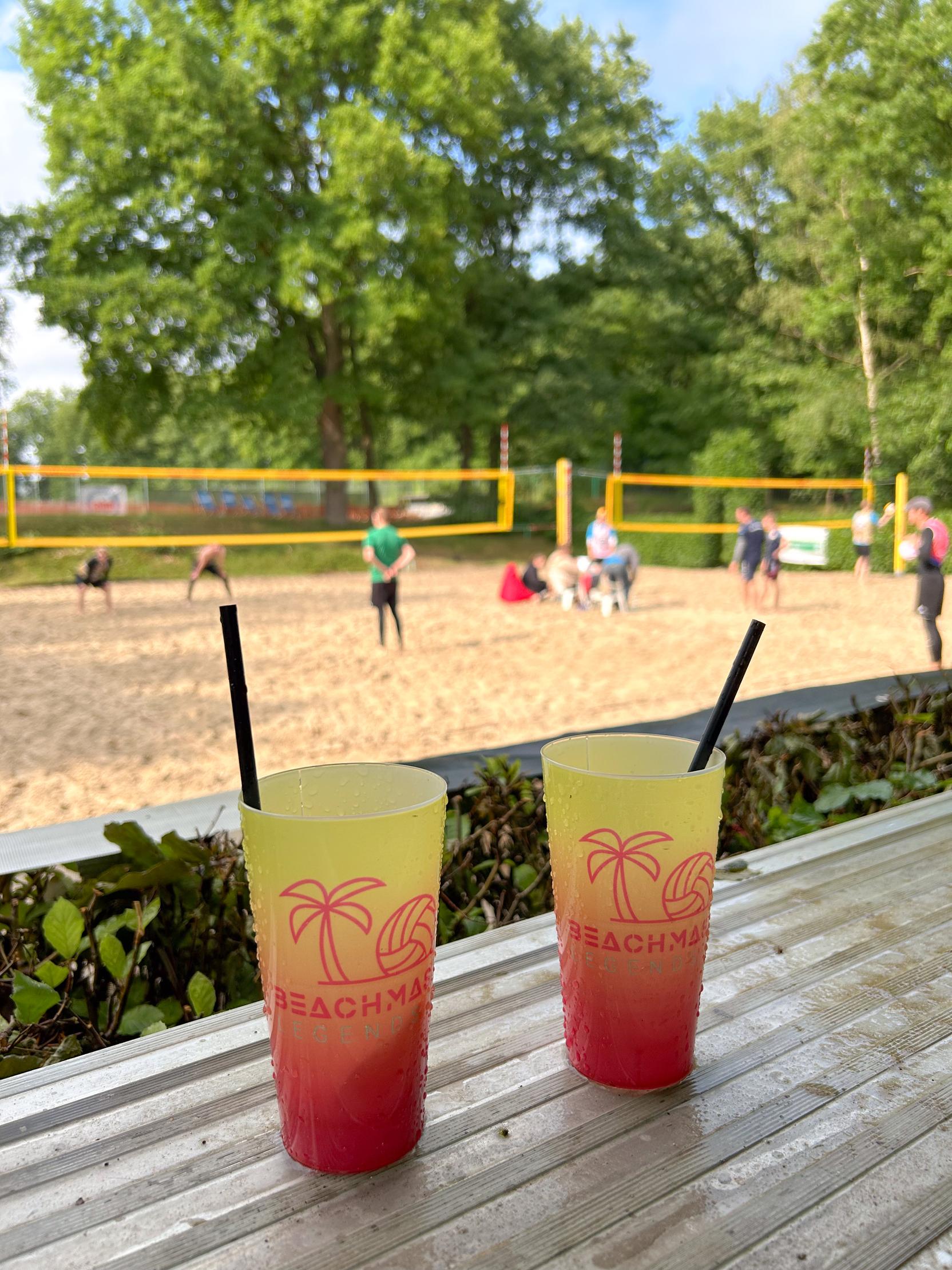 Zwei bunte Cocktails mit Strohhalmen auf einem Tisch, im Hintergrund spielen Menschen Beachvolleyball.
