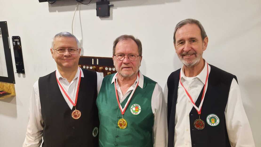 Drei M&auml;nner in festlicher Kleidung mit Medaillen um den Hals stehen nebeneinander vor einer Wand.