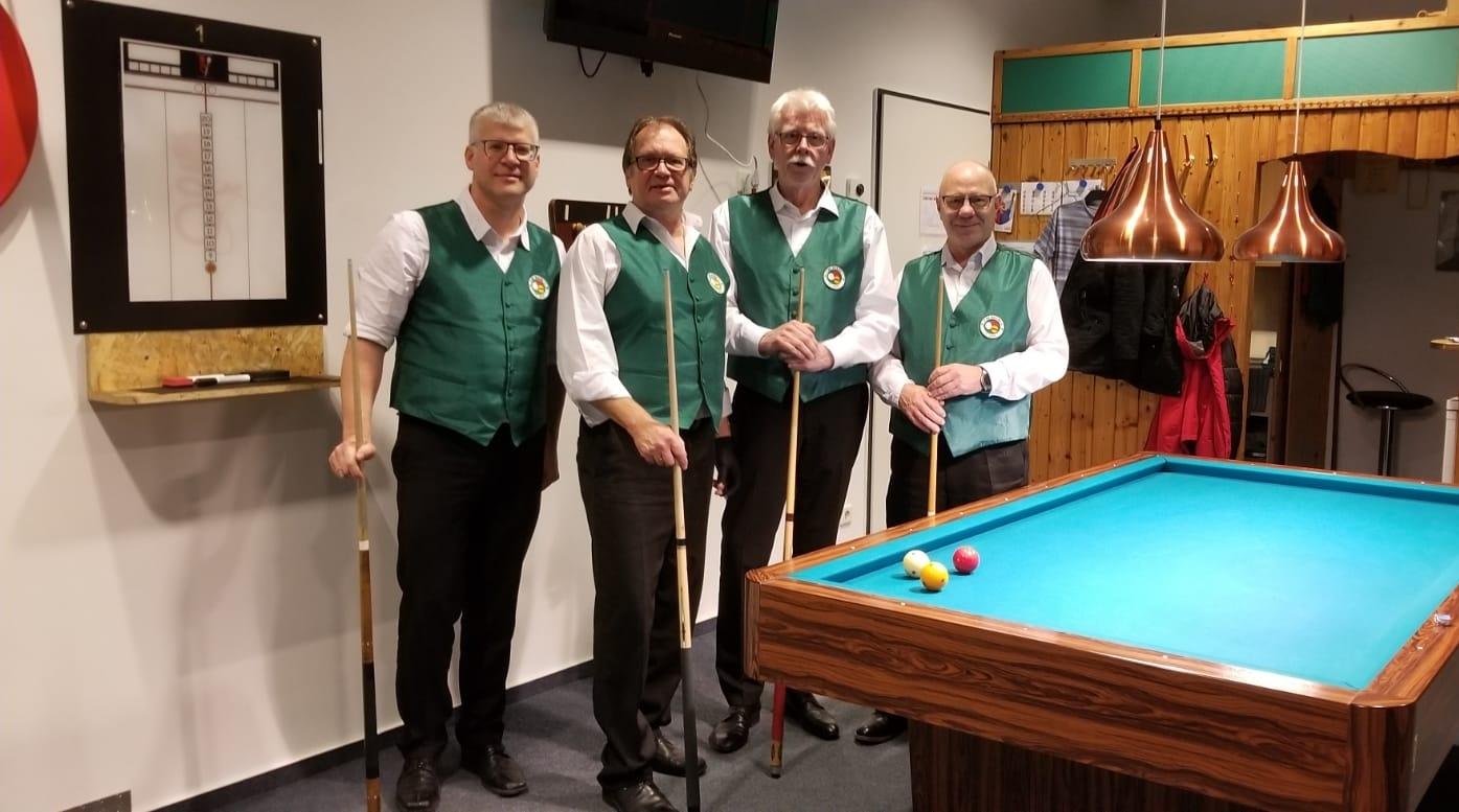Vier M&auml;nner in gr&uuml;nen Westen stehen neben einem Billardtisch mit bunten Kugeln und einem Spielstandsanzeiger im Hintergrund.