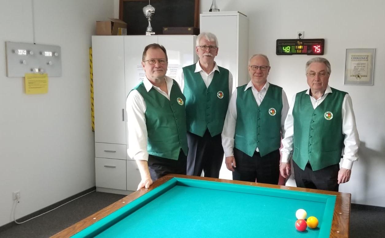 Vier M&auml;nner in gr&uuml;nen Westen stehen vor einem Billardtisch mit drei B&auml;llen, im Hintergrund eine Wand mit Ger&auml;ten und Uhr.