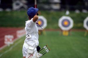 Junge Person in wei&szlig;er Sportbekleidung zielt mit Pfeil und Bogen auf Zielscheiben im Hintergrund.