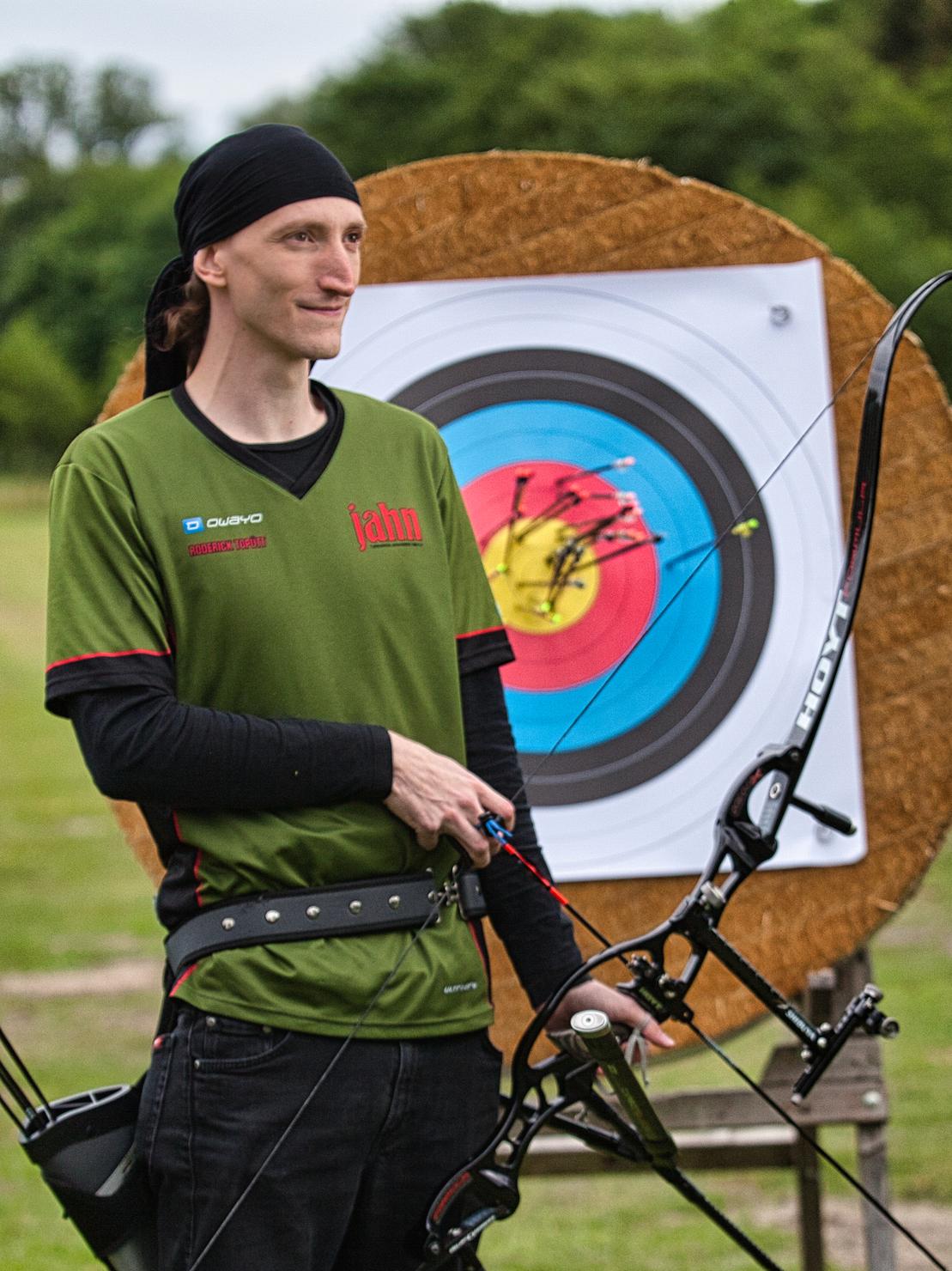 Mann mit Pfeil und Bogen vor einem Ziel mit mehreren Pfeilen in der Mitte, im Hintergrund eine grüne Wiese.