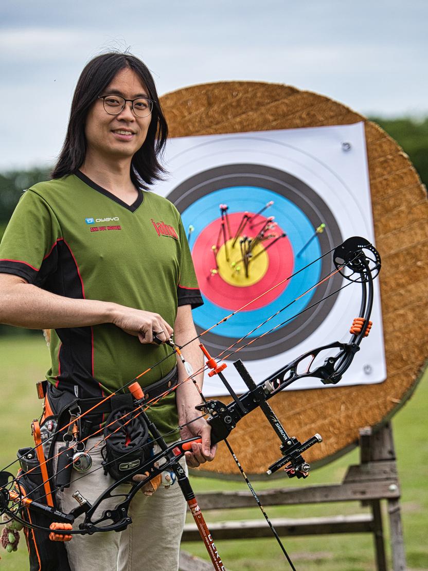 Ein Bogenschütze mit Pfeil und Bogen steht vor einer Zielscheibe mit bunten Trefferzonen auf einem grünen Sportfeld.