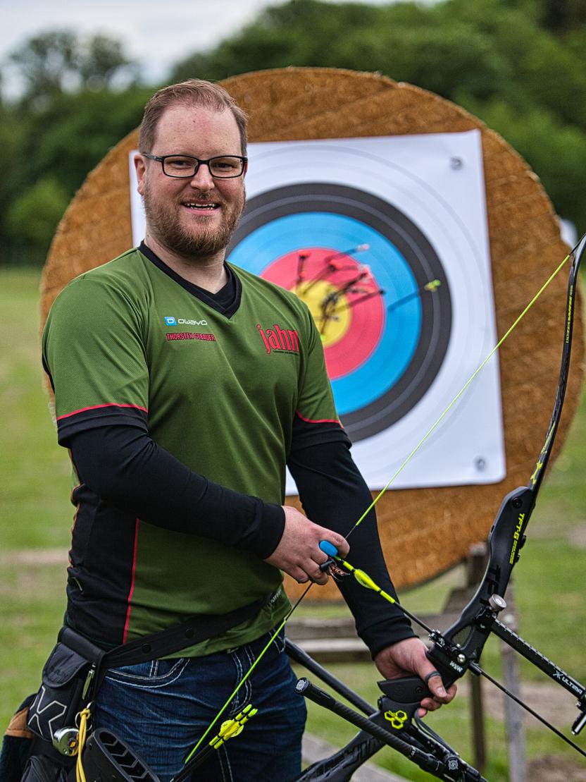 Lächelnder Bogenschütze in Sportkleidung hält einen Bogen vor einer Zielscheibe im Freien.