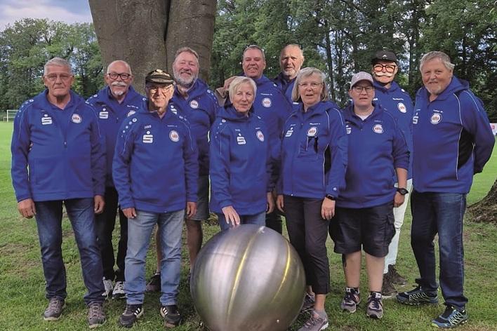 Gruppe von zw&ouml;lf Personen in blauen Shirts steht um eine gro&szlig;e Kugel auf einer Wiese unter einem Baum.