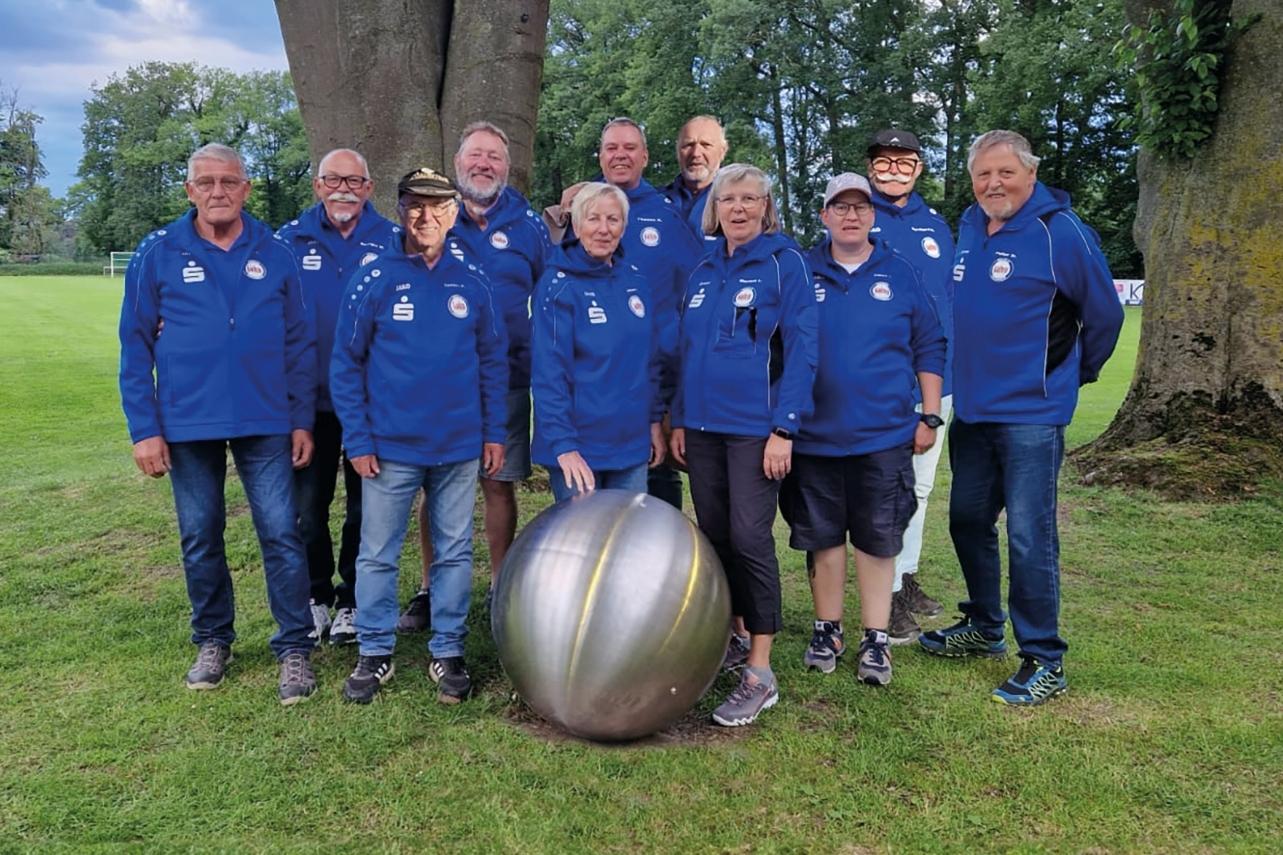 Gruppe von zehn Personen in blauen Jacken steht vor einem Baum, neben einer gro&szlig;en Metallkugel auf Gras.