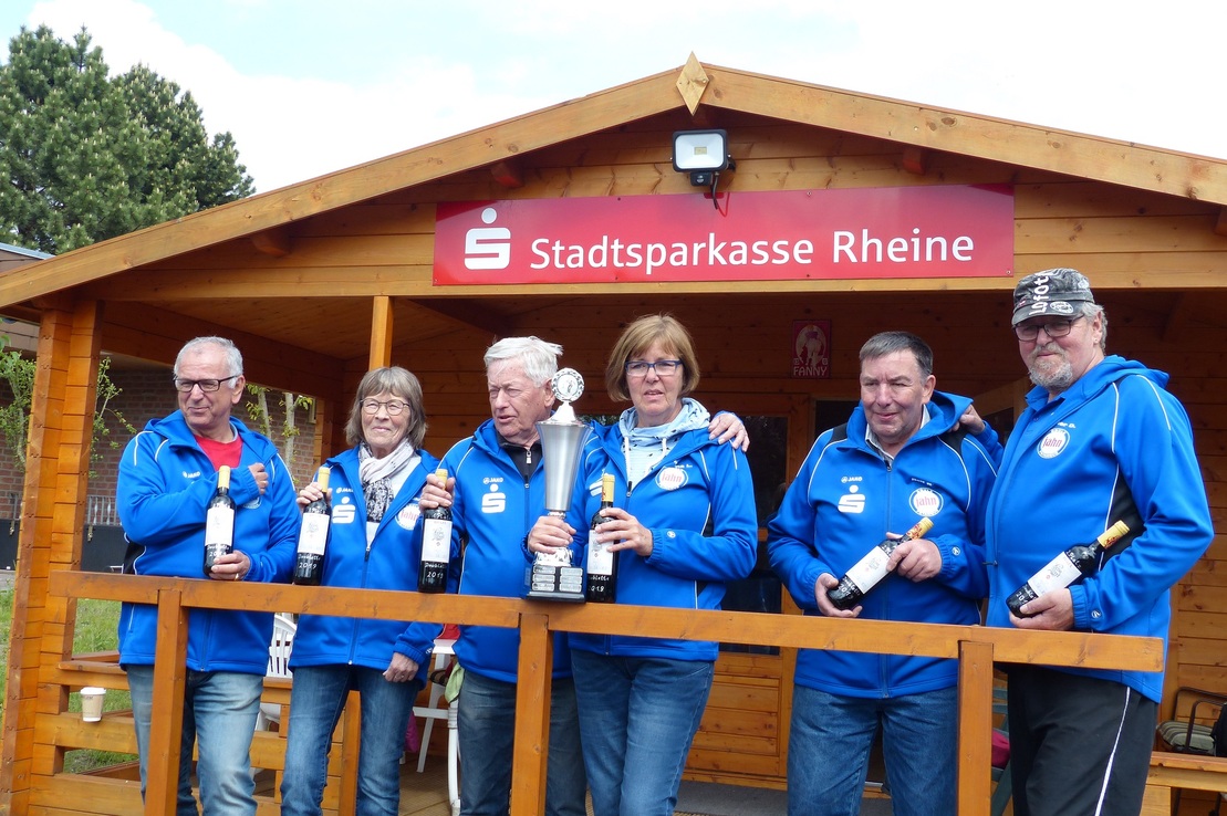 Gruppe von sechs Personen in blauen Sweatshirts stehen vor einem Holzh&auml;uschen mit Troph&auml;en und Praxisflaschen in der Hand.