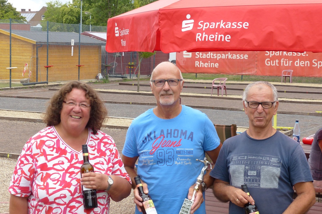 Drei Personen l&auml;cheln am Rande eines Boule-Platzes, halten Flaschen und Pokale unter einem roten Pavillon.