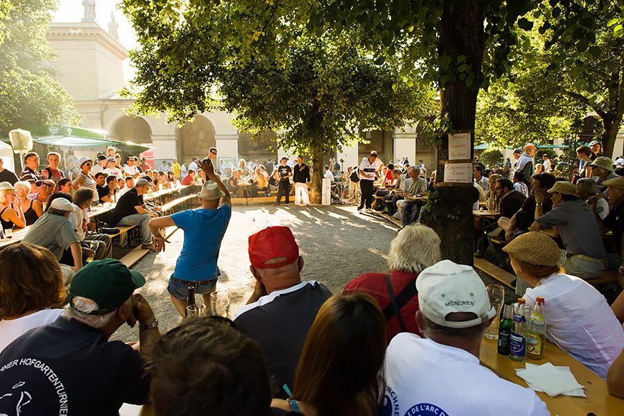Menschen sitzen an Tischen unter B&auml;umen und schauen einem Wettkampf in einem Gartenlokal zu.