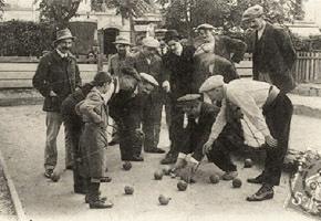 Gruppe von M&auml;nnern und einem Jungen spielt Boule auf einem Platz mit mehreren Kugeln auf dem Boden.