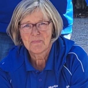 Porträt einer lächelnden Frau mit Brille in blauer Sportkleidung, die auf einer Veranstaltung sitzt.