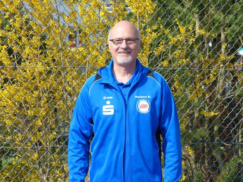 Mann in blauer Sportjacke steht vor einem Zaun mit gelben Blumen im Hintergrund.