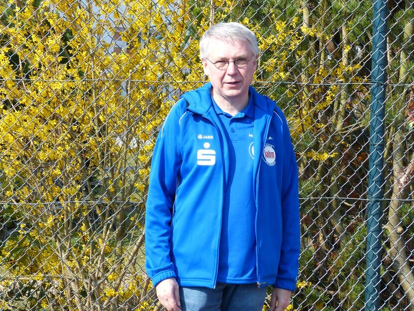 Mann in blauer Jacke steht vor einem Zaun und bl&uuml;henden gelben Str&auml;uchern im Freien.