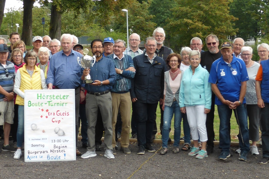 Gruppierung von M&auml;nnern und Frauen bei der Siegerehrung, mit Pokal und Plakat im Freien, umgeben von B&auml;umen.