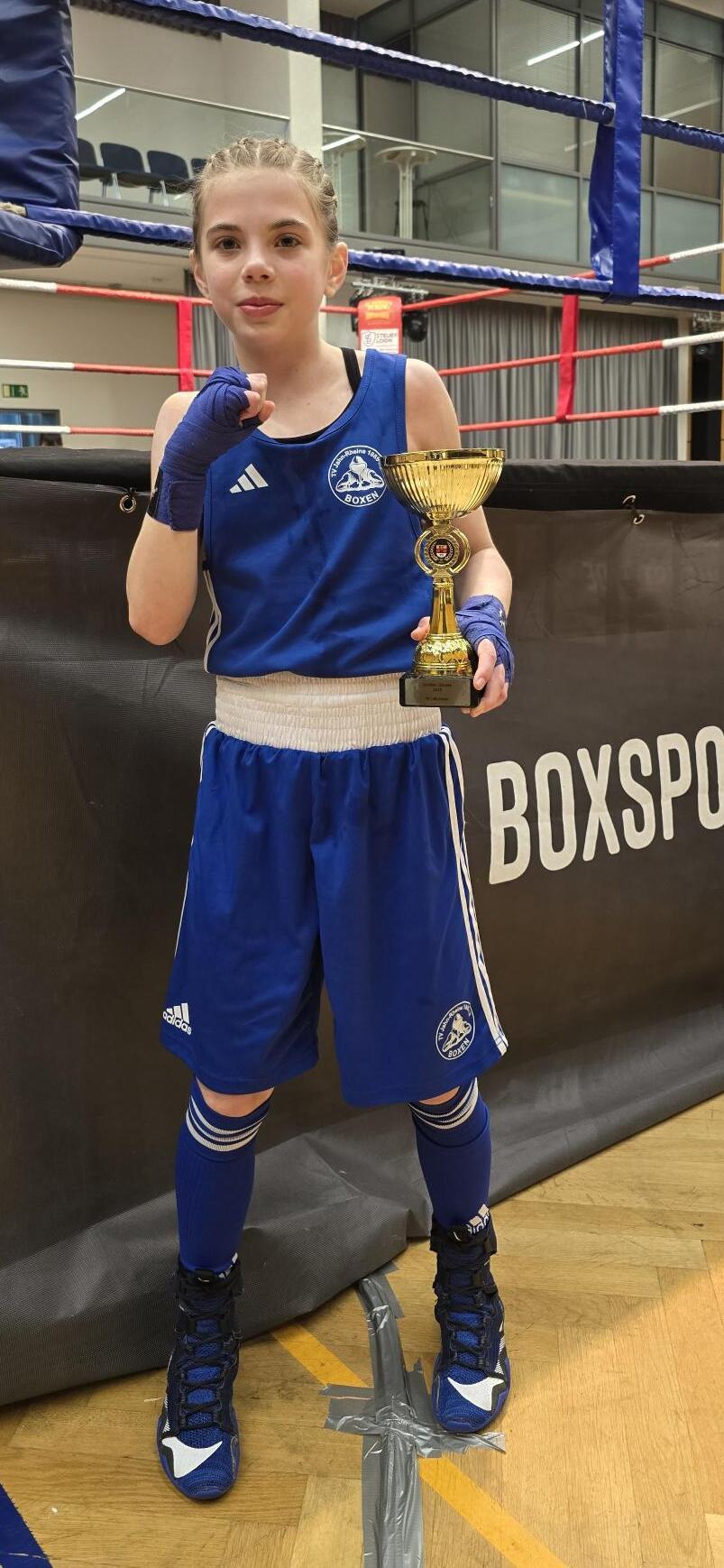 Ein Mädchen in blauer Boxkleidung hält einen Pokal in der Hand und steht vor einem Boxring.