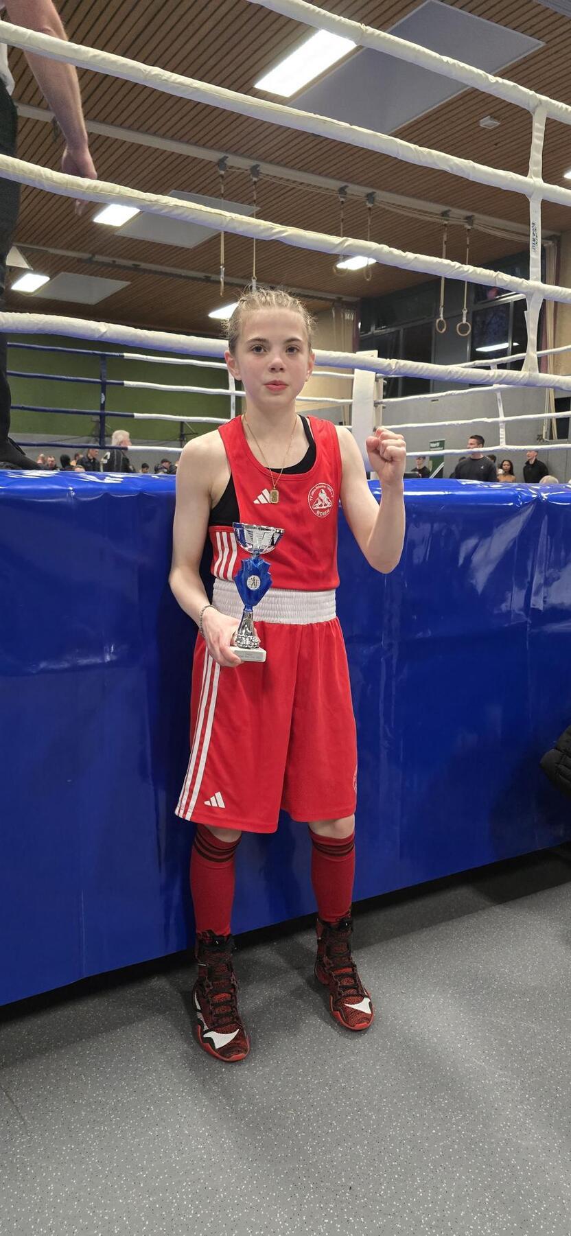 Mädchen im roten Boxanzug hält einen Pokal und zeigt die Siegerfaust vor einem blauen Boxring.