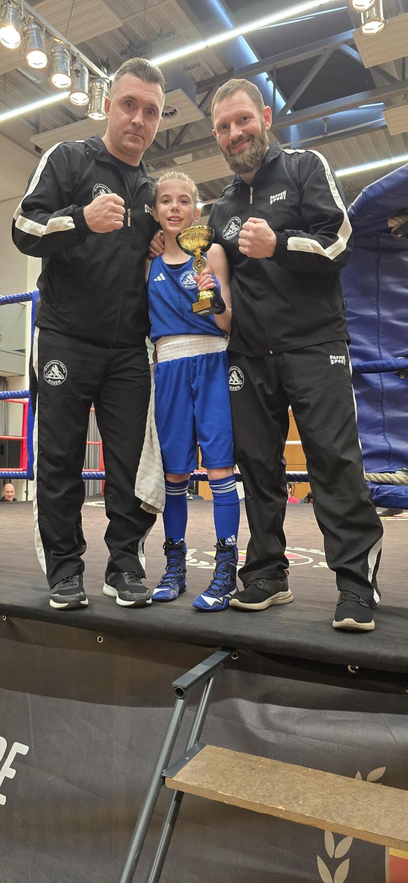 Zwei Trainer und ein Boxerkind halten einen Pokal auf einem Boxring in einer Sporthalle.