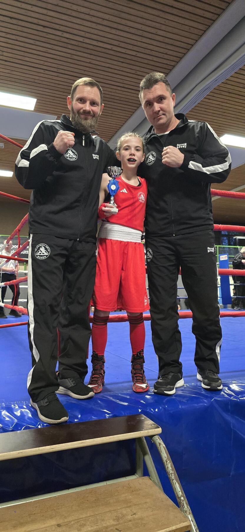 Drei Personen stehen auf einem Ring, die mittlere hält einen Pokal, während die Trainer stolz daneben stehen.
