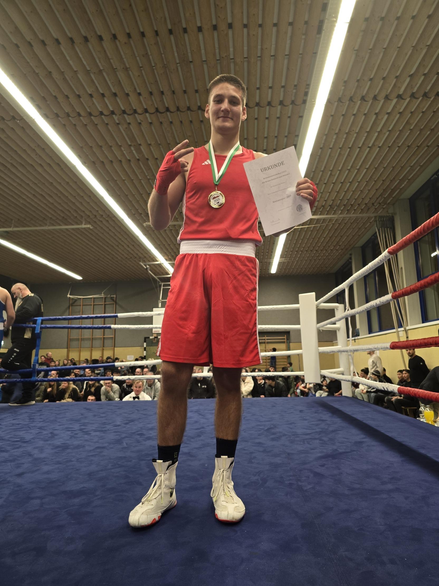 Junger Boxer in roter Kleidung hält eine Urkunde und Medaille auf einem Boxring. Zuschauer im Hintergrund.