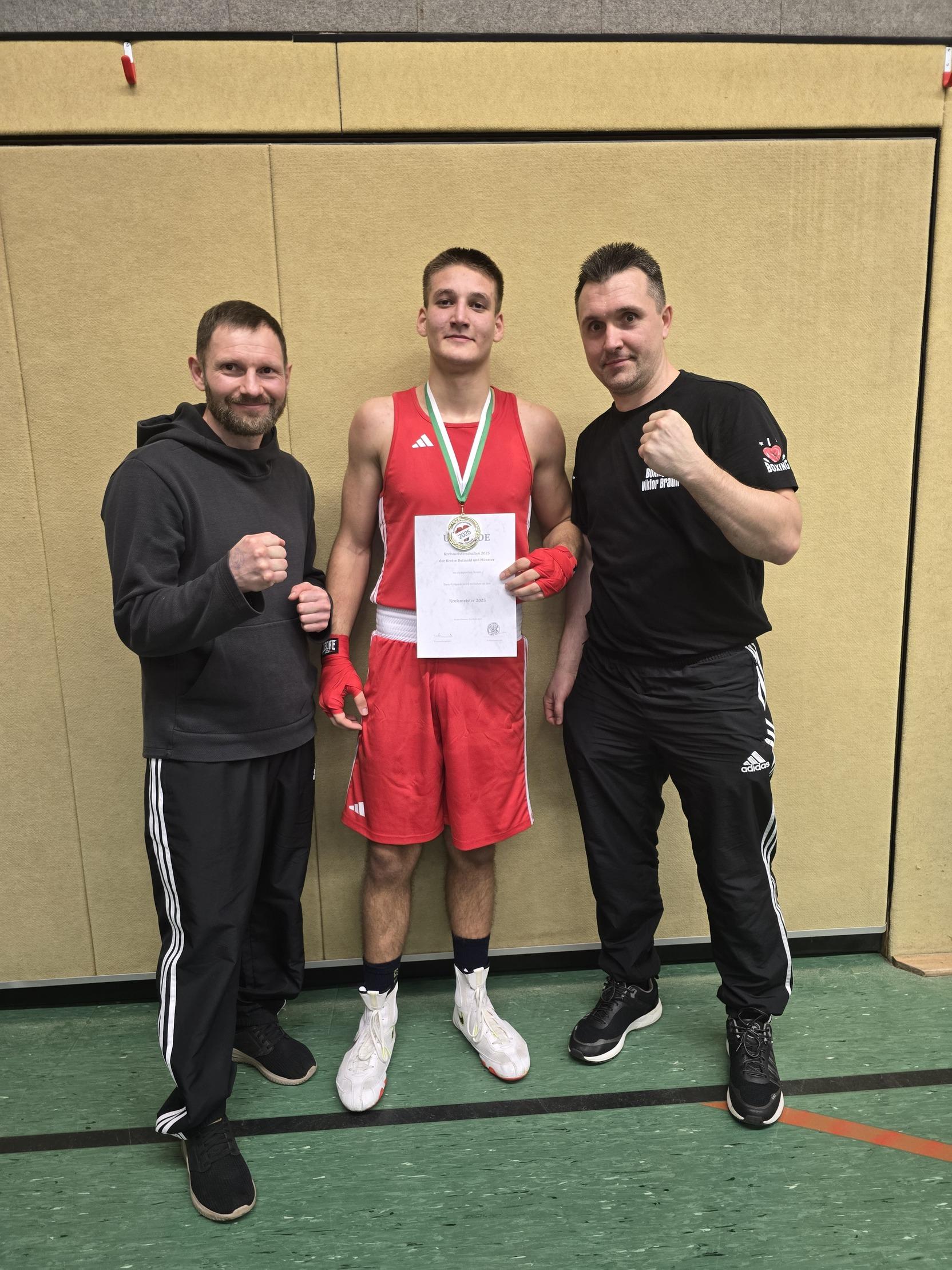Junger Boxer in roter Sportkleidung hält eine Urkunde, umgeben von zwei Trainern in Sportoutfits.