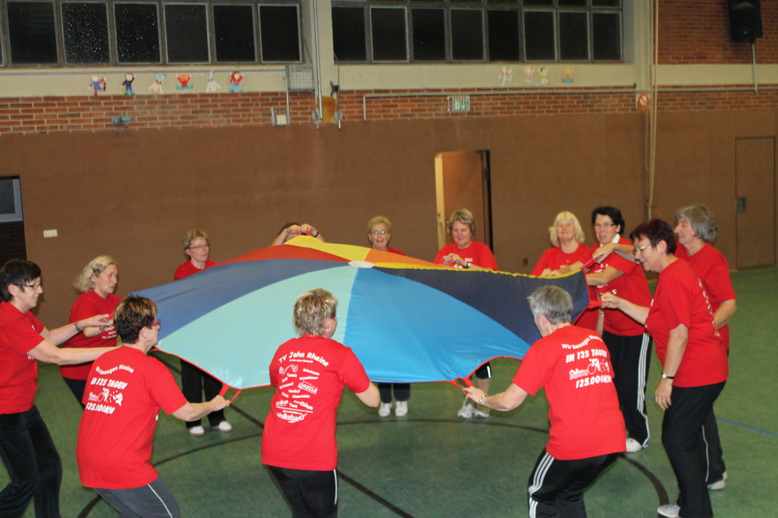 Gruppe von Frauen in roten T-Shirts, die gemeinsam einen bunten Fallschirm in einer Sporthalle halten.